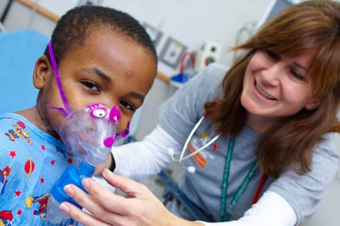 Little boy sitting in hospital bed smiling at camera wearing a nebulizer with nurse assisting him.