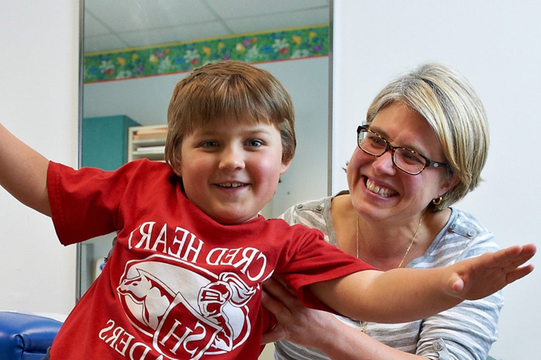 Boy doing airplane arms with his mom behind him smiling.