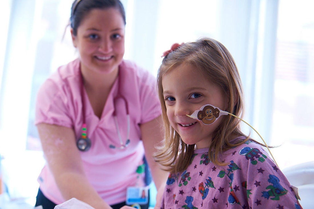 Young girl wearing pink patterned hospital gown sits on bed with tube in her nose smiling, a nurse stands behind her smiling.