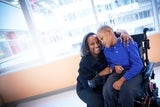 In the hospital, a boy in a wheelchair, wearing blue, is embraced by a smiling older woman, offering loving support