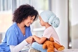 A girl in a blue headscarf holds a teddy bear, smiling at a nurse with curly hair in a hospital room.