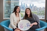 Two women sitting holding up a ceramic memory plate with  handprints made by a family where the child is now deceased