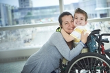 Boy in wheelchair wearing stripe shirt smiling and hugging his mother in hospital setting.