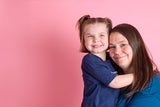 Young girl hugging her mother, both in blue shirts on pink background.