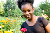 Young girl in black tee shirt smiling and standing in a field of flowers with a red daisy in hand.