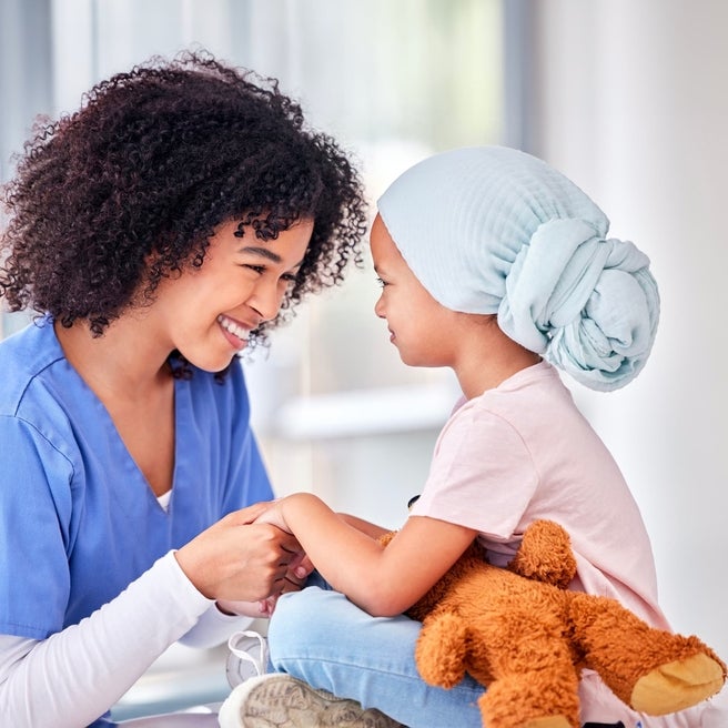 A girl in a blue headscarf holds a teddy bear, smiling at a nurse with curly hair in a hospital room.