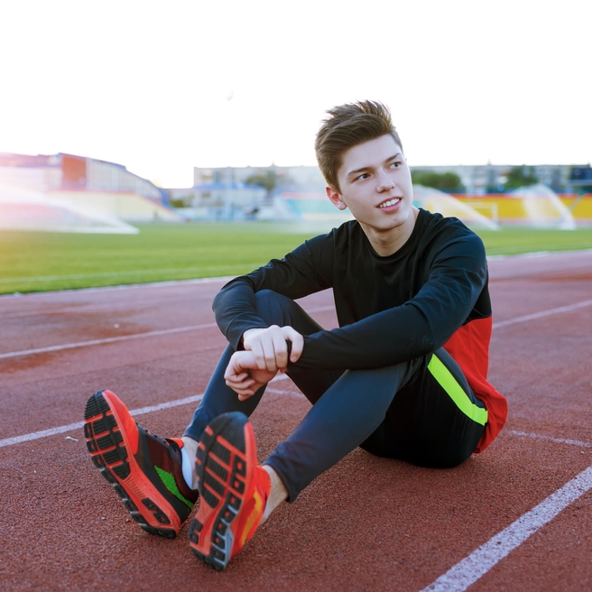 Older teen boy sitting on a track wearing cleats looking up.