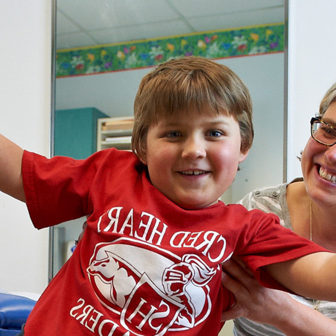 Boy doing airplane arms with his mom behind him smiling.