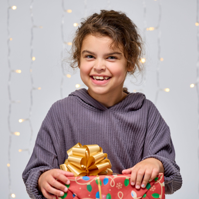 Smiling girl holding a holiday gift with twinkling lights in background.