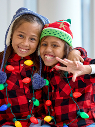 Two school-age girls wearing holiday lights and winter clothes, smiling and holding up peace signs