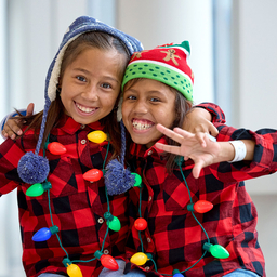 Two school-age girls wearing holiday lights and winter clothes, smiling and holding up peace signs