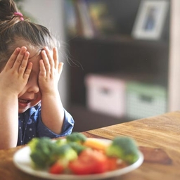 Young girl with hands over eyes sitting in front of a plate of food