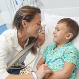 Parent smiling and holding the child’s hand while he’s laying down in a hospital bed.